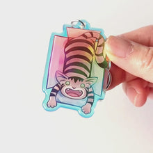 Load and play video in Gallery viewer, A clear, blue tinted holographic acrylic keychain featuring a character from Rainbow of Crazy Comics: a smiling tabby cat lounges in a cardboard box.
