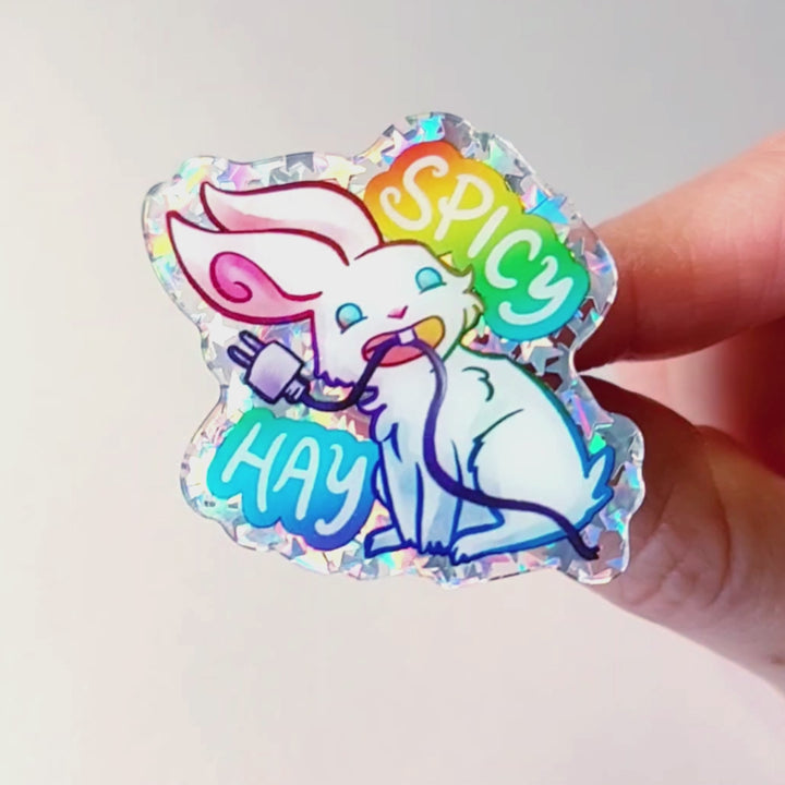 sparkly acrylic pin of a smiling white rabbit holding a charging cable in her mouth, with the words "SPICY HAY in rainbow colours.