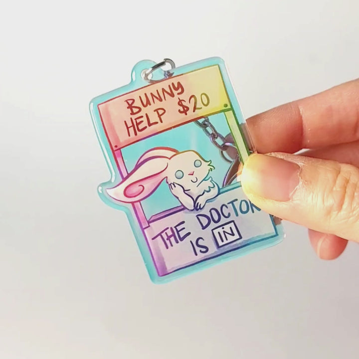 A clear, rainbow holographic acrylic keychain featuring a character from Rainbow of a Crazy Comics: a smiling white rabbit at a homemade booth that says "bunny help $20" at the top, and "the doctor is in" at the bottom. 
