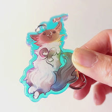 Load and play video in Gallery viewer, A clear, rainbow holographic acrylic keychain featuring a character from Rainbow of Crazy Comics: a smiling fluffy ragdoll cat, lying on her back, with a tiny black-capped chickadee, the bird perched on the cat's ear.
