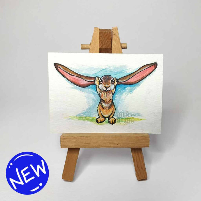an aceo painting, featuring a brown rabbit holding up it's extra long ears, painted against a splash of blue and green. the painting sits on a tiny wood easel
