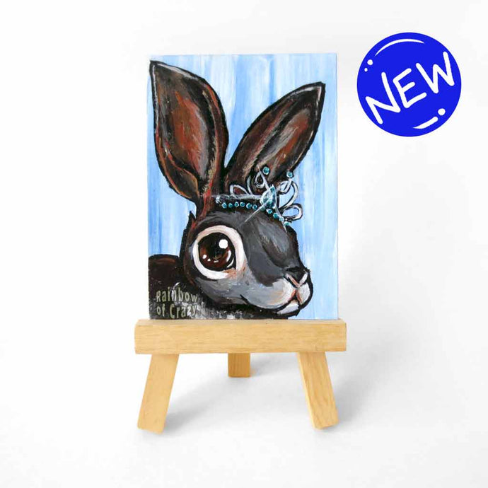 an aceo sized painting featuring a close up of a rabbit wearing a sparkling tiara, background painted a light blue. the painting sits on a tiny wood easel