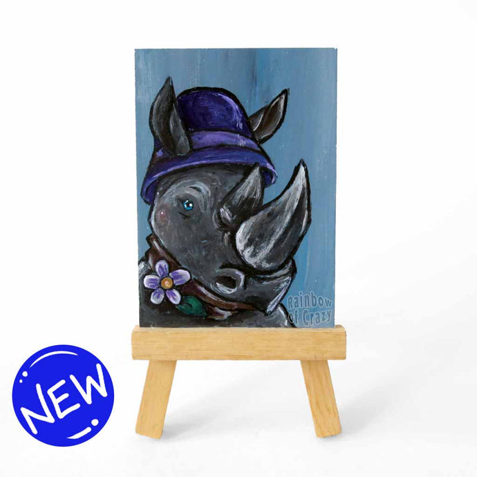 aceo sized painting of a rhino wearing a purple cloche hat and purple flower on a scarf. the painting sits on a small wood easel