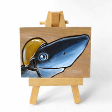 Load image into Gallery viewer, an aceo sized painting featuring a smiling shark, wearing a gold jockey helmet. the painting rests on a small wood easel
