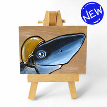 Load image into Gallery viewer, an aceo sized painting featuring a smiling shark, wearing a gold jockey helmet. the painting rests on a small wood easel
