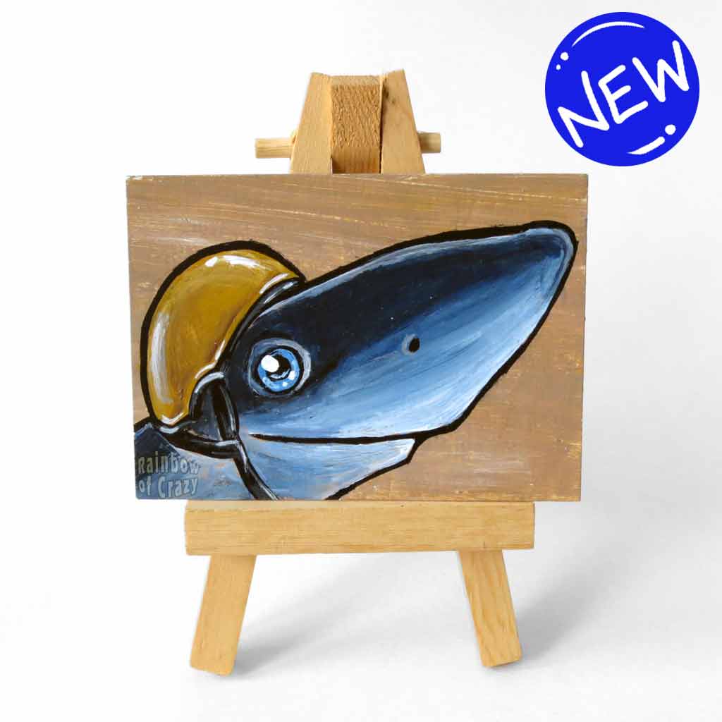 an aceo sized painting featuring a smiling shark, wearing a gold jockey helmet. the painting rests on a small wood easel