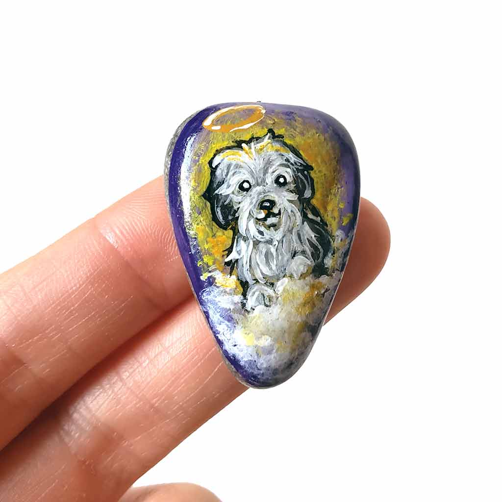 a beach stone hand painted with a sheepdog, as an angel with a halo, sitting on clouds against a purple and yellow sky. available as a keepsake or pendant necklace