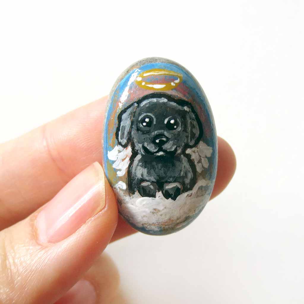 A small beach stone, hand painted with a portrait of a black shih tzu dog as an angel sitting in the clouds. Available as a keepsake or necklace