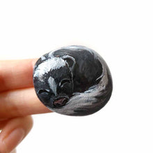 Load image into Gallery viewer, a small beach stone, painted to look like a sleeping baby skunk
