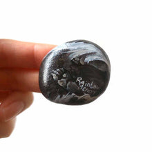 Load image into Gallery viewer, a small beach stone, painted to look like a sleeping baby skunk. the underside shows tiny feet, and is signed with RainbowofCrazy
