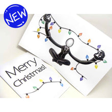 Load image into Gallery viewer, a greeting card with outside text: &quot;Merry Christmas!&quot; with a string of lights beneath / Inside features an illustration of Slender Man sitting cross-legged, arms out stretched, holding a long line of christmas lights
