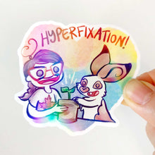 Load image into Gallery viewer, Heart shaped sticker with the word "HYPERFIXATION!" above a Comic Jo and an English Spot rabbit, both smiling as they tend to a potted plant.

