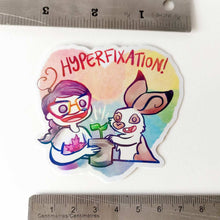 Load image into Gallery viewer, Heart shaped sticker with the word "HYPERFIXATION!" above a Comic Jo and an English Spot rabbit, both smiling as they tend to a potted plant.

