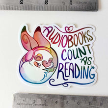 Load image into Gallery viewer, holographic sticker with a smiling rabbit wearing headphones. the sticker reads, "Audiobooks count as reading"
