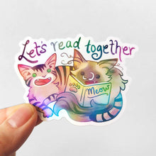 Load image into Gallery viewer, a sticker with a tabby cat next to a ragdoll cat that's holding a book titled "Meow". At the top of the sticker, it says, "Let's read together".
