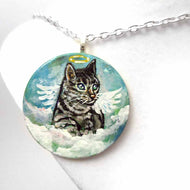 a small wood disc, hand painted with the portrait of a gray tabby cat, as an angel with wings and halo, sitting on clouds against a blue sky. this painting is available as a keepsake or pendant necklace