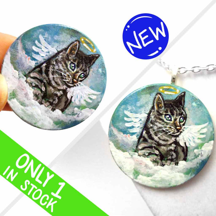 a small wood disc, hand painted with the portrait of a gray tabby cat, as an angel with wings and halo, sitting on clouds against a blue sky. this painting is available as a keepsake or pendant necklace