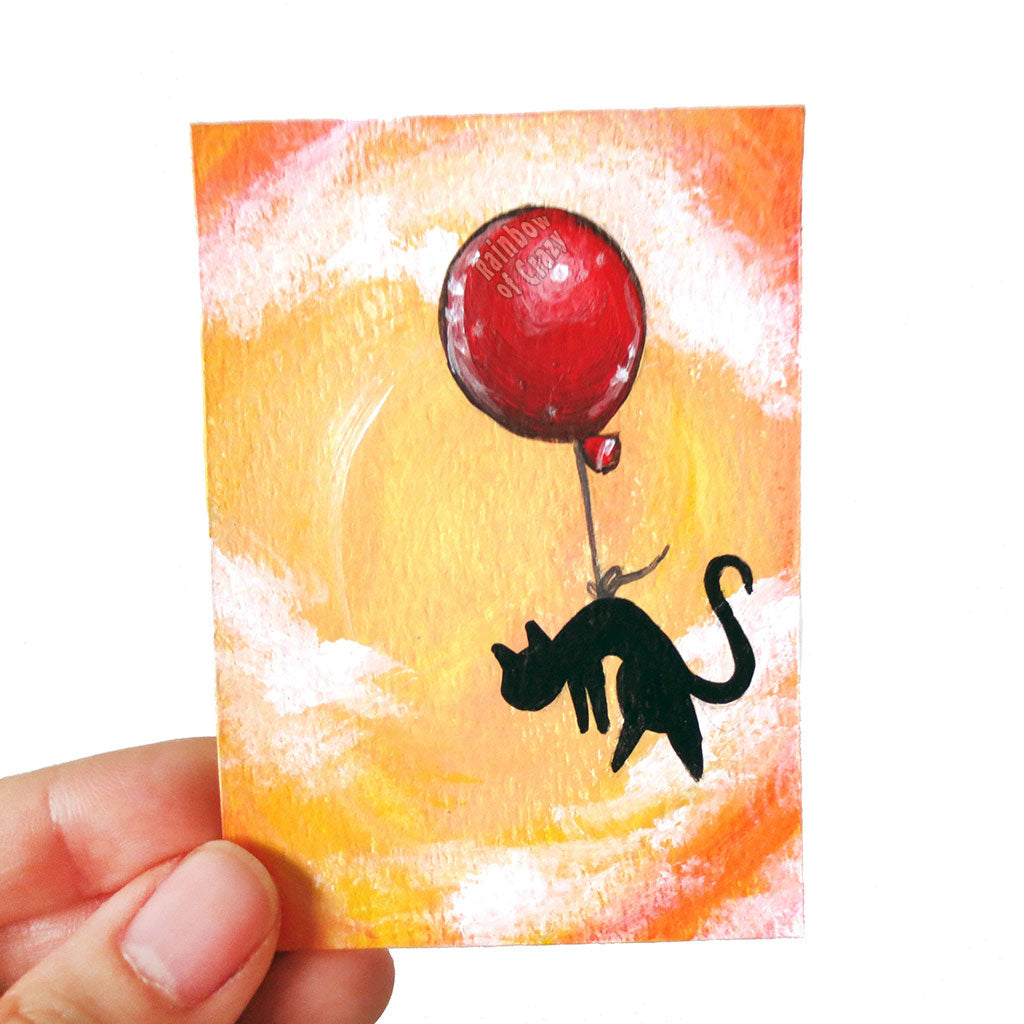 A hand holding a small ACEO size painting of a black cat hanging from a red balloon, floating through a cloudy orange sky.