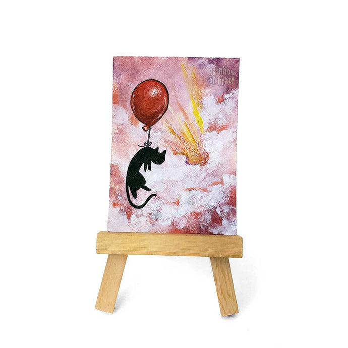 An original ACEO painting, on an easel, of a black cat with a red balloon, floating  through the clouds in the sky, painted with reds and purples. 