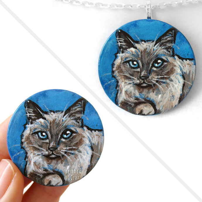 A circle wooden disc, with cat art of a Birman cat with blue eyes, with a blue background. Available as either a wood keepsake or pendant necklace