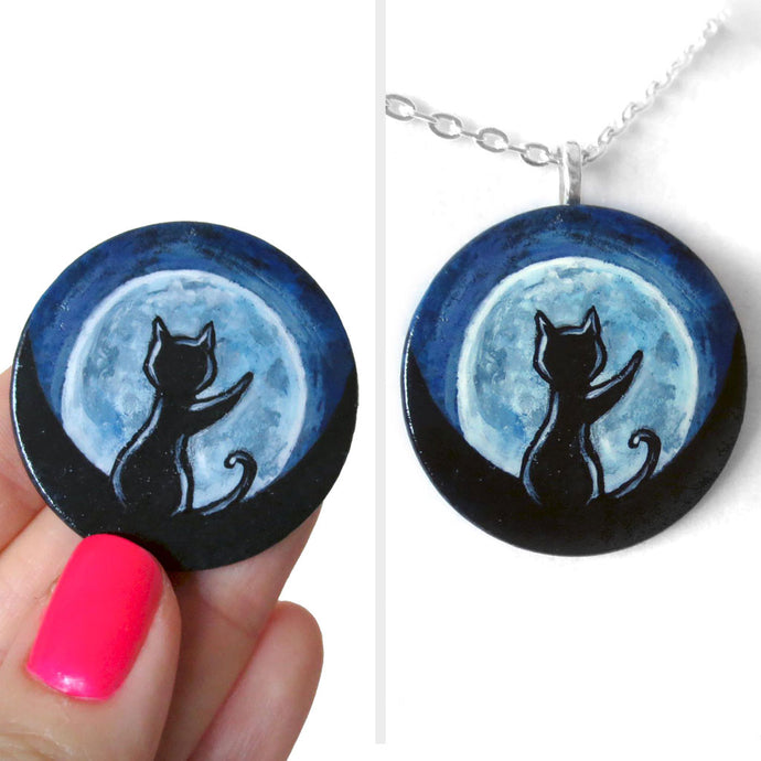 A lightweight wooden disc features art of the silhouette of a black cat, with a front paw reaching up, sitting in front of a big glowing full moon. Available as a keepsake or necklace