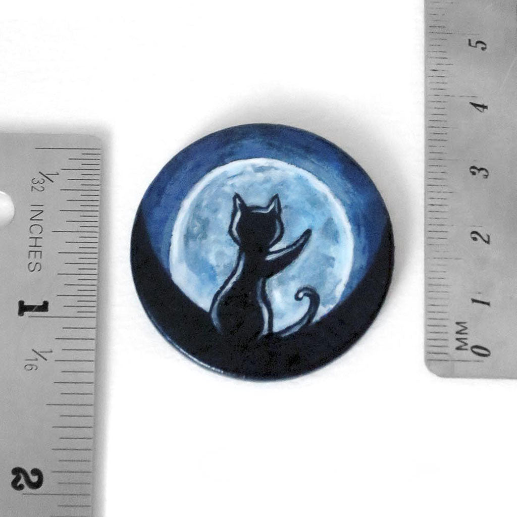 A wooden circle painted with a black cat in front of the full moon, placed next to two rules to show its size: 1 1/2 inches or 3.8 cm across