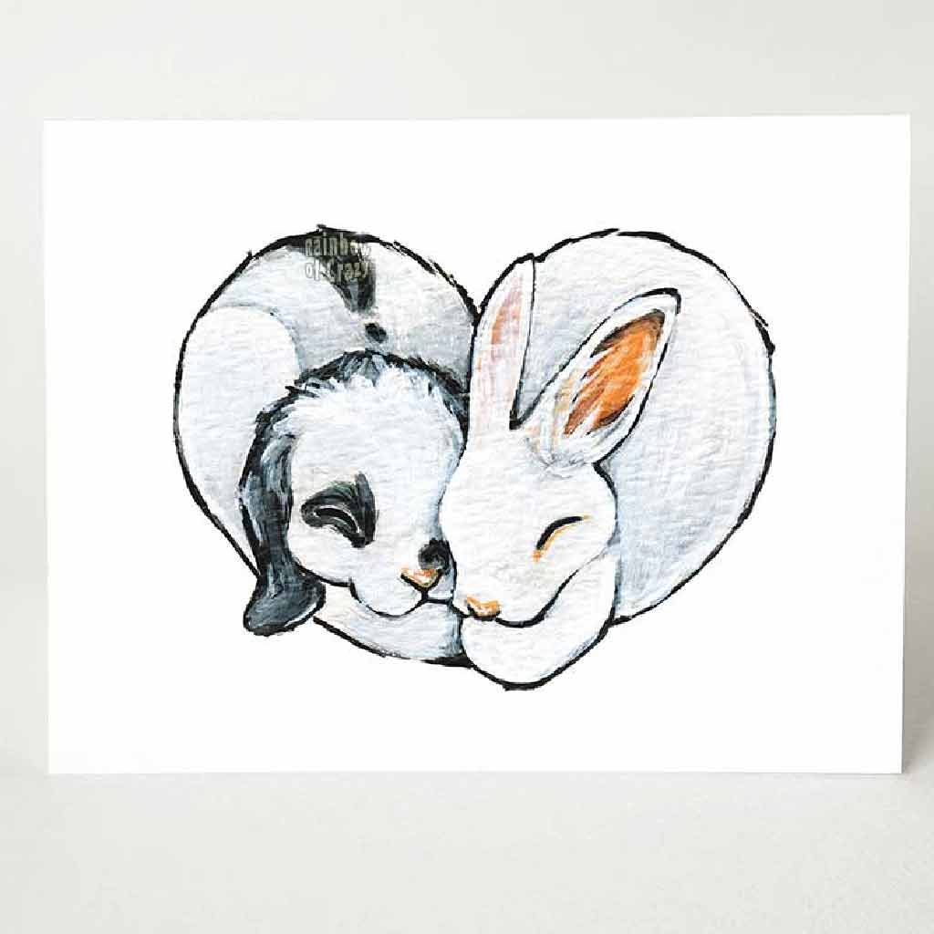 A greeting card, printed with art of two rabbits (a white and black mini lop, and a white Polish), cuddled together in the shape of a heart.