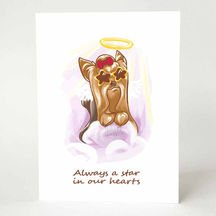 A greeting card with an illustration of a Yorkshire Terrier as an angel, wearing star shaped sunglasses and a red bow on its head, it sits on purple clouds. The card reads, 