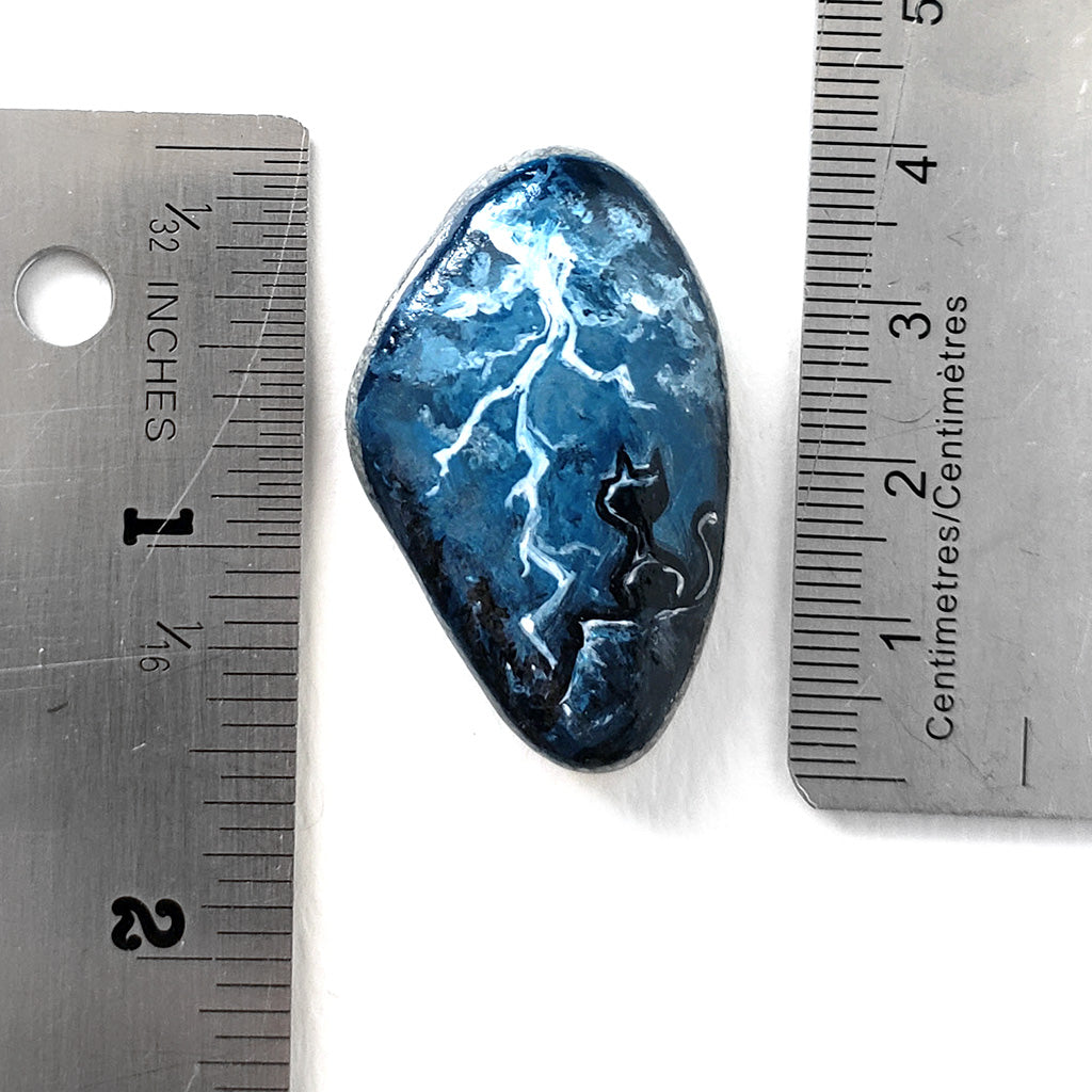 a beach rock with cat art: a black cat on a cliff, looking out into the night sky filled with lightning. the stone is next to two rulers to show its size: 1 9/16