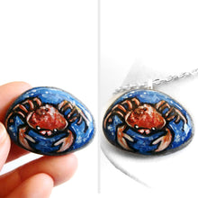 Load image into Gallery viewer, a small beach rock, hand painted with zodiac art: an orange and red crab, the sign of cancer, surrounded by blue water. available as a stone keepsake or pendant necklace
