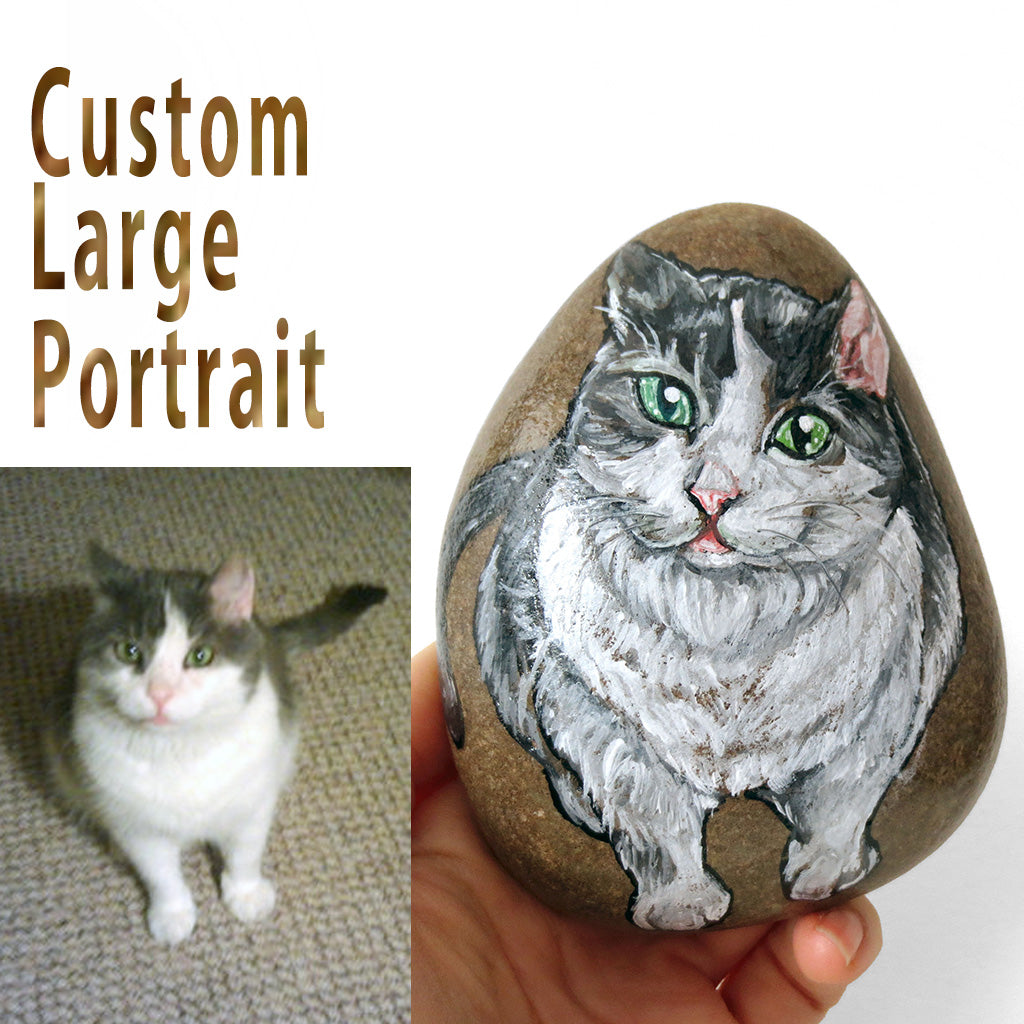 A large beach stone, hand painted with a custom portrait of a grey and white cat with green eyes