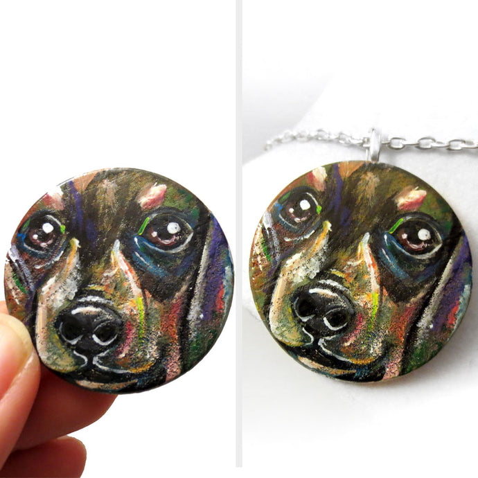 A round wood pendant is hand painted with a portrait of a dacshund dog, and is available as either a keepsake or a pendant necklace