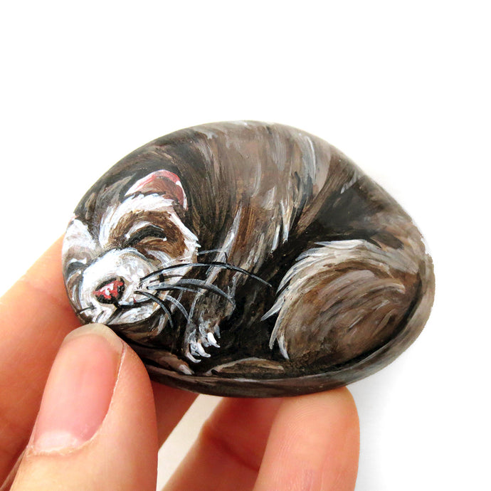 A beach stone has been hand painted with the portrait of a brown ferret sleeping.