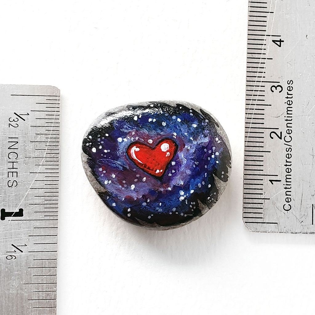 a beach stone, painted with a heart in a starry galaxy sky, next to two rulers to show its size. it measures 1 3/16