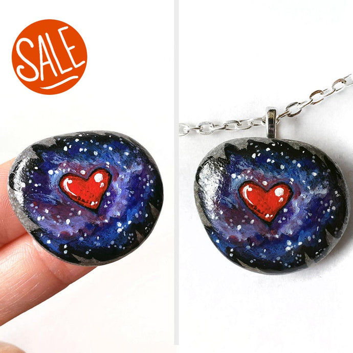 a beach stone has been hand painted with a heart surrounded by outer space and stars, and hand crafted as either a keepsake stone or pendant necklace