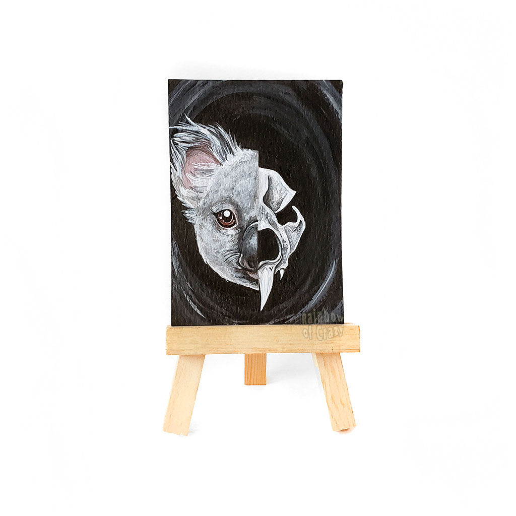 a small easel with a miniature painting that is ACEO sized (2.5 inches by 3.5 inches), of a split portrait: the left side is painted with a koala, and the right side is painted with a stylized koala skull. 