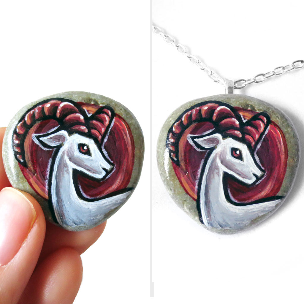 a beach rock painted with the zodiac art of aries: a white ram with dark red horns. this stone is available as a keepsake or pendant necklace