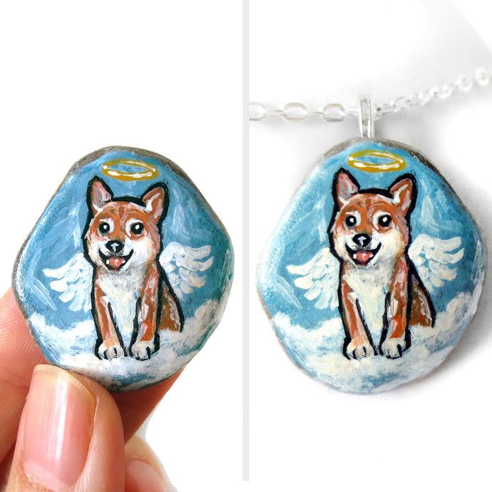 a smooth beach stone, hand painted with a portrait of a shiba inu dog as an angel, sitting on clouds against a light blue sky. available as a keepsake or pendant necklace