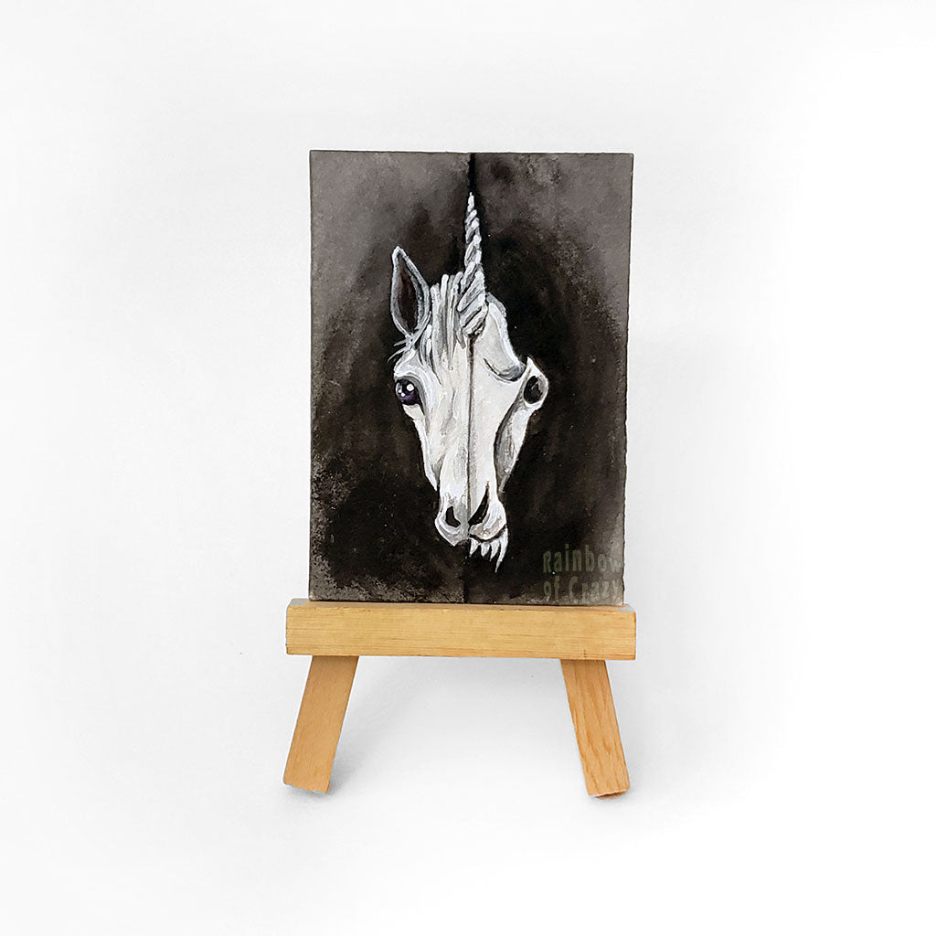an easel with an ACEO sized miniature painting (2.5 inches by 3.5 inches) of a split image: the left side is a white unicron and the right side is a unicorn skull with sharp fangs