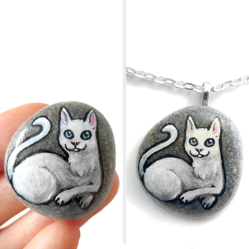 A small beach stone hand painted with a portrait of a white cat with blue eyes, available as a stone or pendant necklace.