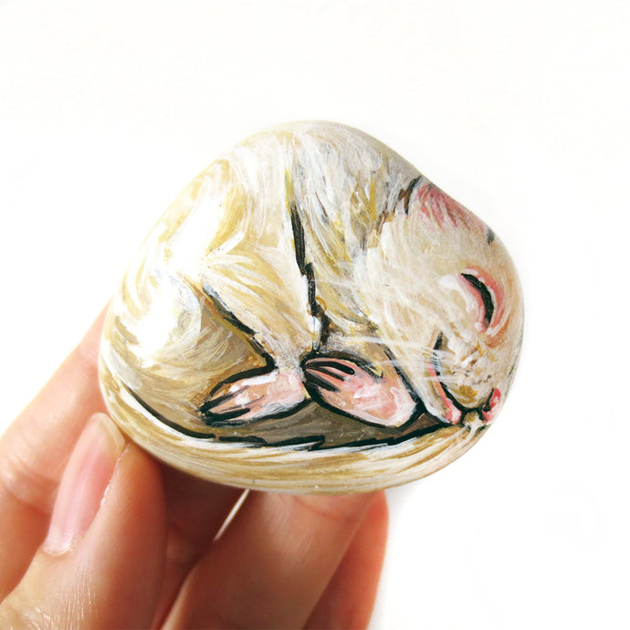 A hand holding a small beach stone painted with a portrait of an albino ferret sleeping.