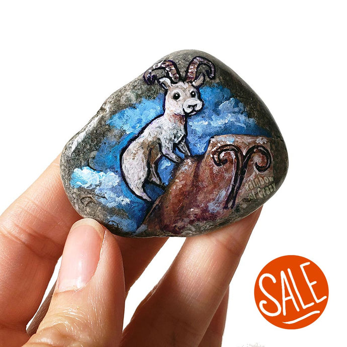 Rock art featuring a painting of a ram on a mountain, in front of a cloudy blue sky. The symbol for Aries is painted on the bottom.