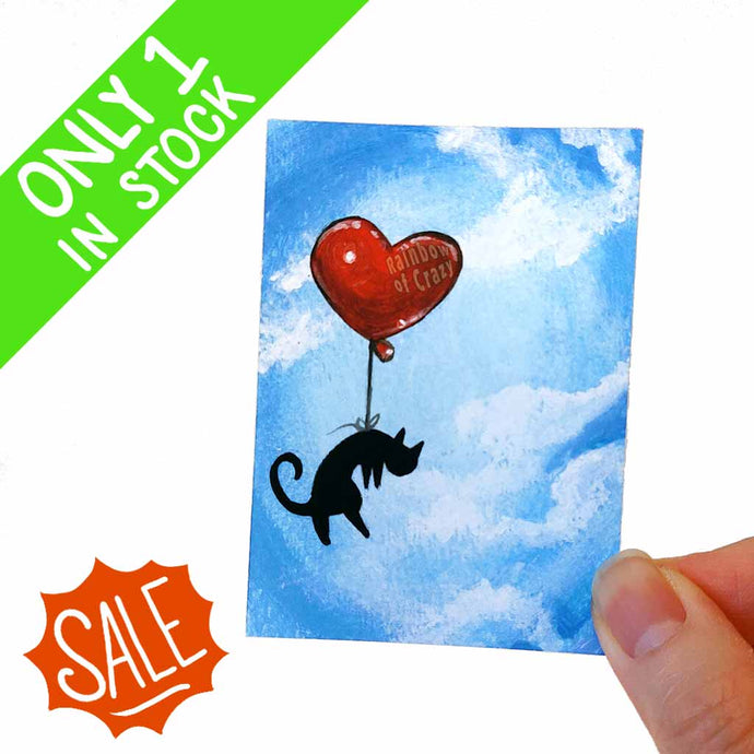 an aceo art print features a black cat floating through the cloudy blue skies, thanks to a big heart shaped red balloon tied around its waist