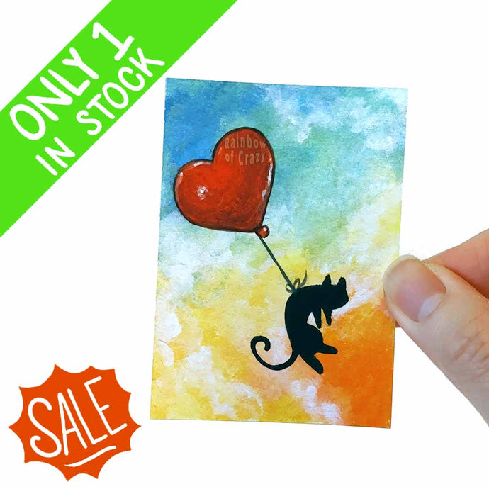 an aceo print of a black cat floating through a cloudy sunset sky with a red heart shaped balloon tied around its waist