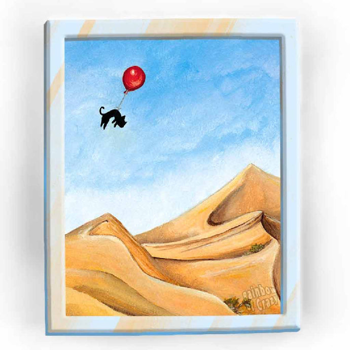 An art print with an illustration of a black cat floating with a red balloon, soaring over the Sarah desert