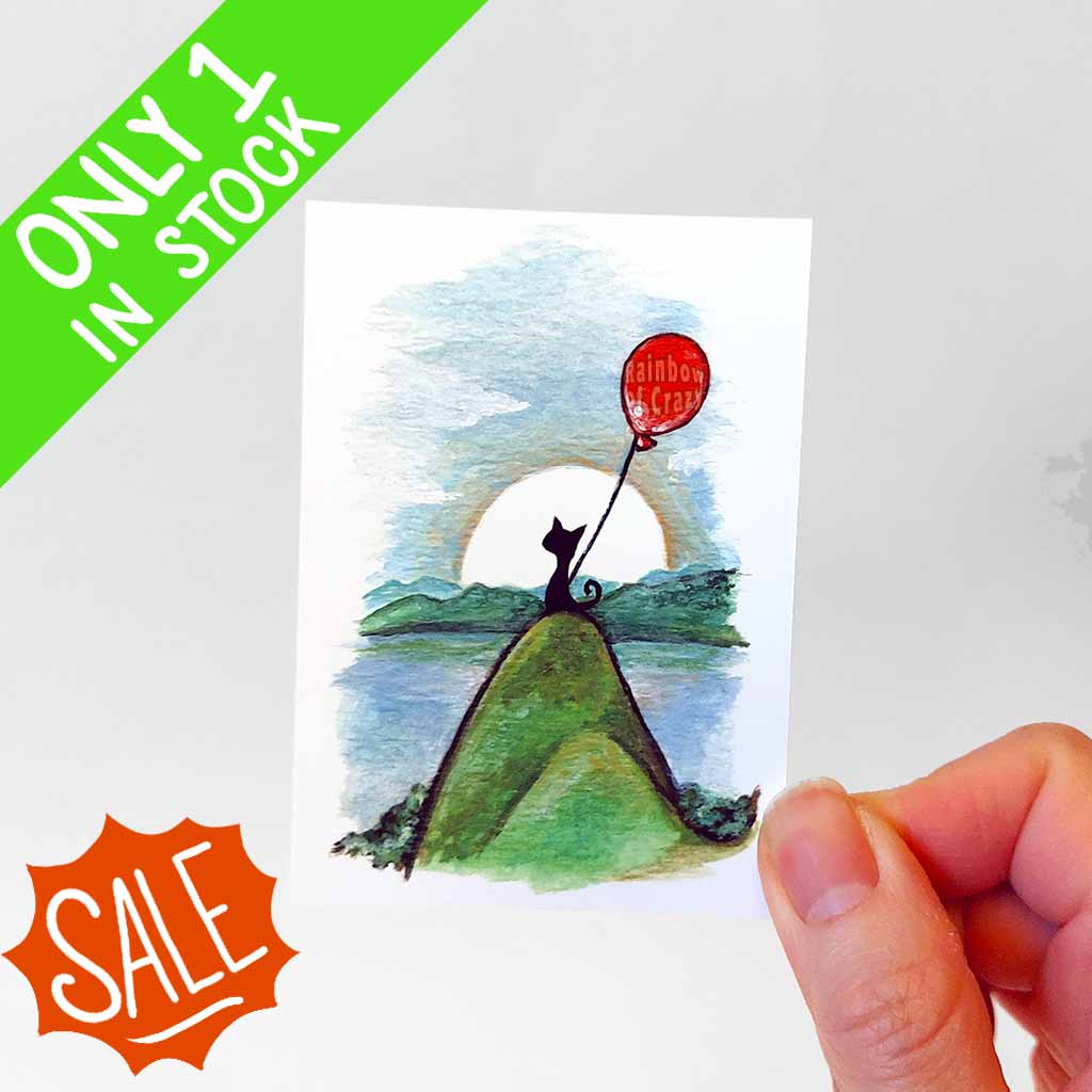 an aceo print that features a black cat sitting on a hill overlooking mountains and blue waters. The kitty holds a shiny red balloon and the sun shines from behind