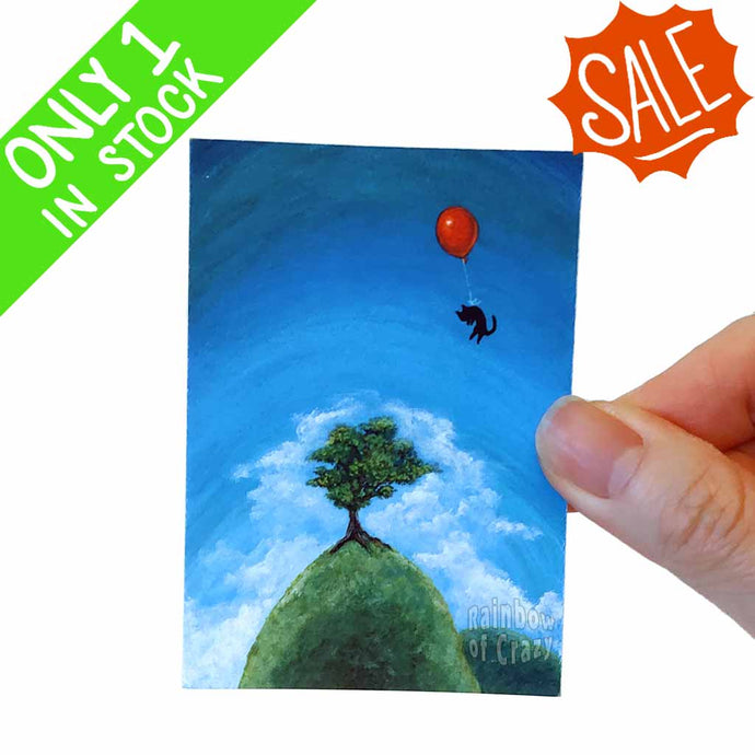 an aceo art print featuring a tiny black cat following through a blue sky with a red balloon tied to its waist. it floats over a tree on a tall hill 