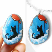 Load image into Gallery viewer, a small beach stone, hand painted with a little black cat, with a red balloon, sitting on clouds against a blue sky. available as a keepsake or pendant necklace
