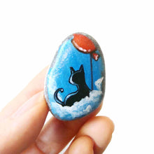 Load image into Gallery viewer, a small beach stone, hand painted with a little black cat, with a red balloon, sitting on clouds against a blue sky. available as a keepsake or pendant necklace
