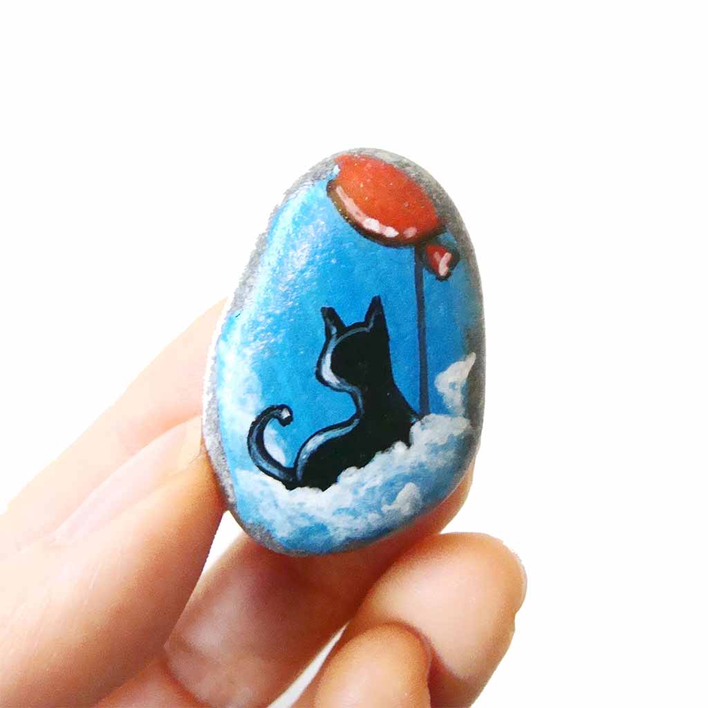 a small beach stone, hand painted with a little black cat, with a red balloon, sitting on clouds against a blue sky. available as a keepsake or pendant necklace